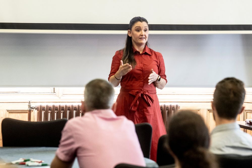 Dark haired female wearing a dark russet red shirt dress presenting to a seated workshop audience in front of a large overhead screen in a light blue room.